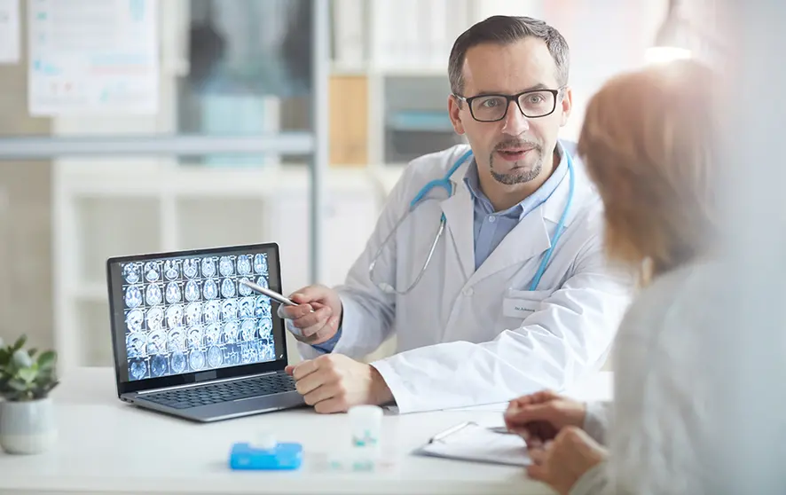 Male doctor explaining the illness to his patient and pointing at laptop with x-ray images while they sitting at the table at office