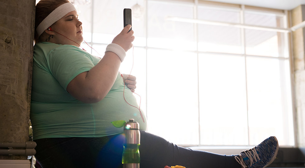 Full length side view portrait of pensive fat woman sitting on mat and using smartphone while taking break in workout